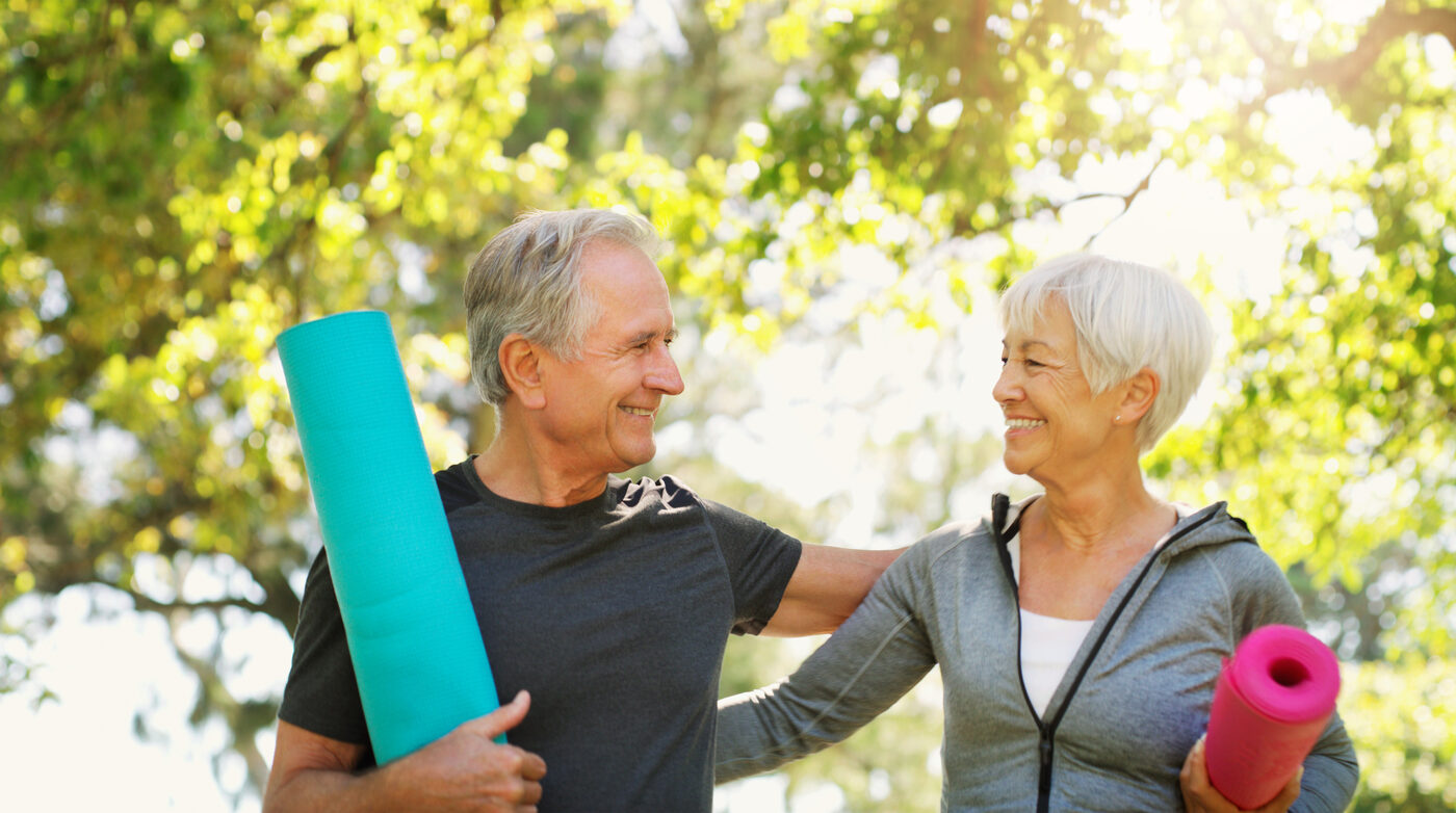 An older couple smiling warmly at each other outdoors, holding yoga mats